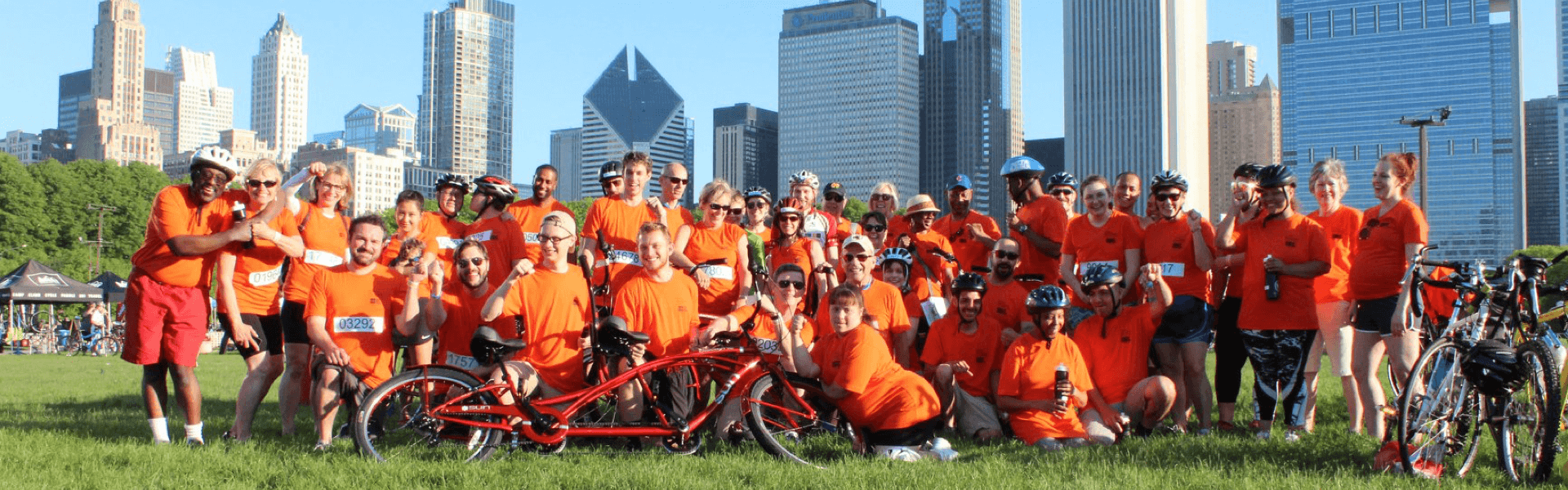 Large group photo outdoors with cityline in the background while wearing orange "Bike the Ride" t-shirts and posing with bikes
