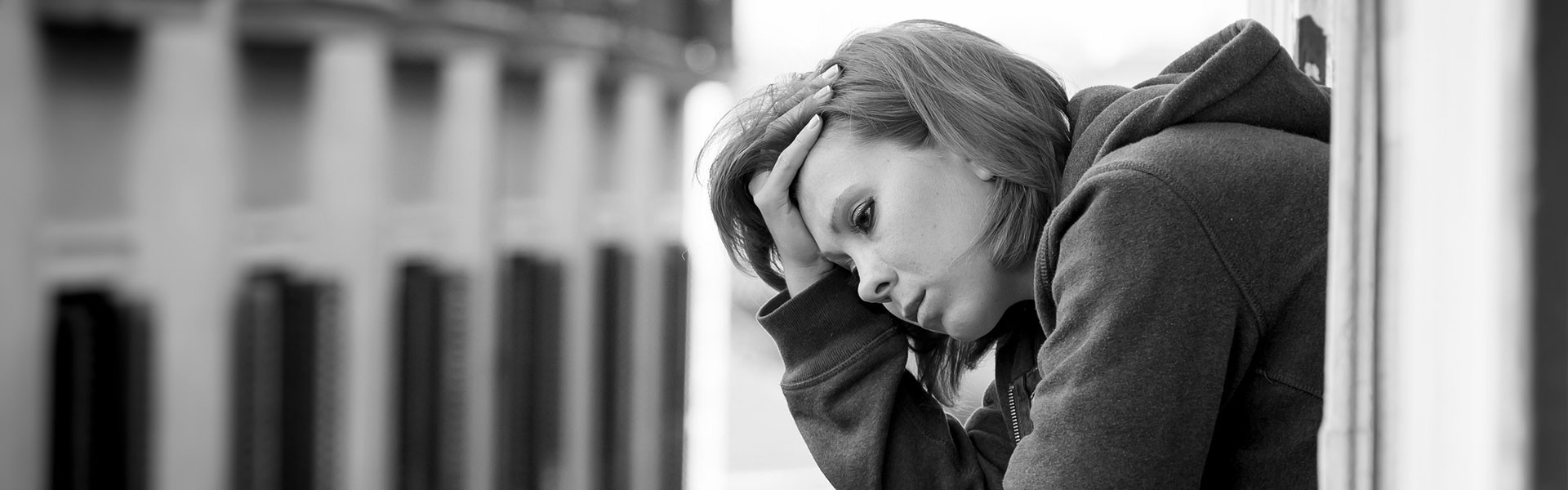 The black and white image shows a person leaning against a wall, looking down with a somber expression. They are wearing a hooded sweatshirt, with the hood down. Their hand is resting on the side of their head, fingers threaded through their hair. The background is blurred, suggesting an urban setting with tall buildings. The grayscale tones emphasize the solemn mood of the scene.