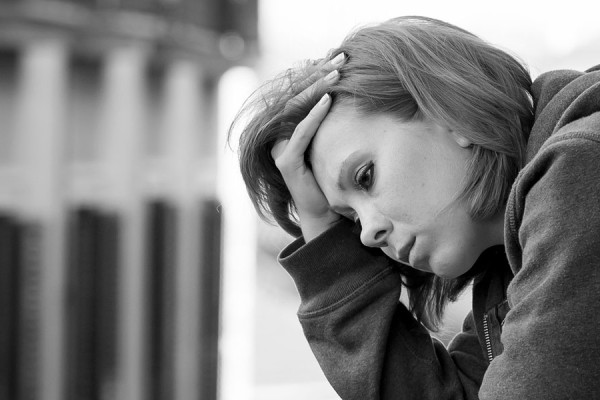 The black and white image shows a person leaning against a wall, looking down with a somber expression. They are wearing a hooded sweatshirt, with the hood down. Their hand is resting on the side of their head, fingers threaded through their hair. The background is blurred, suggesting an urban setting with tall buildings. The grayscale tones emphasize the solemn mood of the scene.